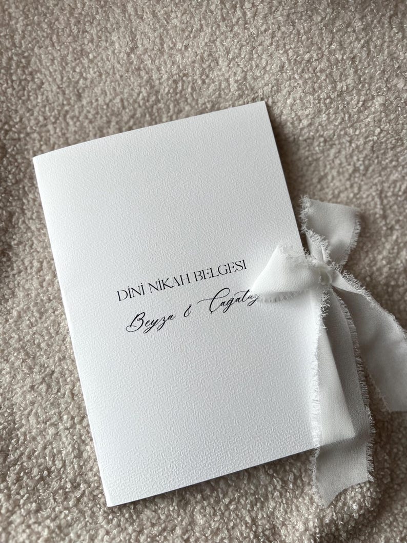 Pode incluir: Um convite branco com o texto "DINI NIKAH BELGESI" e os nomes "Beyza & Cagatay" em escrita cursiva. Uma fita branca est&aacute; amarrada ao redor do convite.