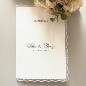 Può includere: Un registro di famiglia color crema intitolato STAMMBUCH con i nomi Selin & Deniz e FAMILIE GÜVEN stampati in inchiostro nero. La copertina presenta un delicato bordo in pizzo bianco ed è arricchita da un bouquet di fiori artificiali.
