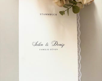 Könnte beinhalten: Ein cremefarbenes Stammbuch mit der Aufschrift STAMMBUCH, den Namen Selin & Deniz sowie FAMILIE GÜVEN in schwarzer Schrift. Der Einband ist mit einer feinen weißen Spitzenborte verziert und wird von einem künstlichen Blumenstrauß ergänzt.