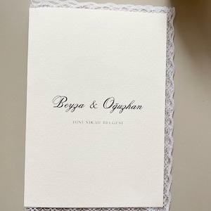Könnte beinhalten: Eine rechteckige, cremeweiße Karte mit den Namen Beyza & Oğuzhan in schwarzer Schreibschrift und dem darunter gedruckten Text DINI NIKAH BELGESI. Die Karte ist an der Ober-, Unter- und rechten Seite mit einer zarten weißen Spitzenborte verziert.