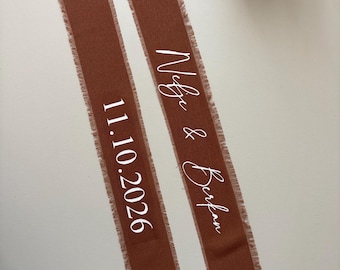 Può includere: Due lunghe e strette strisce di tessuto color ruggine con bordi sfrangiati. Una striscia presenta il testo stampato in bianco 11.10.2026, mentre l'altra mostra il testo stampato in bianco Nefje & Berkan in un carattere corsivo.
