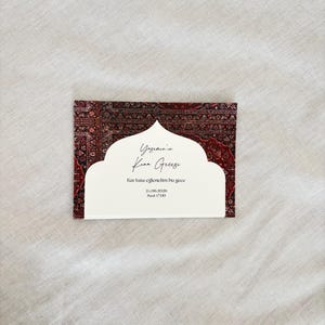 May include: A rectangular invitation card with a white cutout design and text. The card features a patterned red and brown background reminiscent of a rug. The text is in a script font.