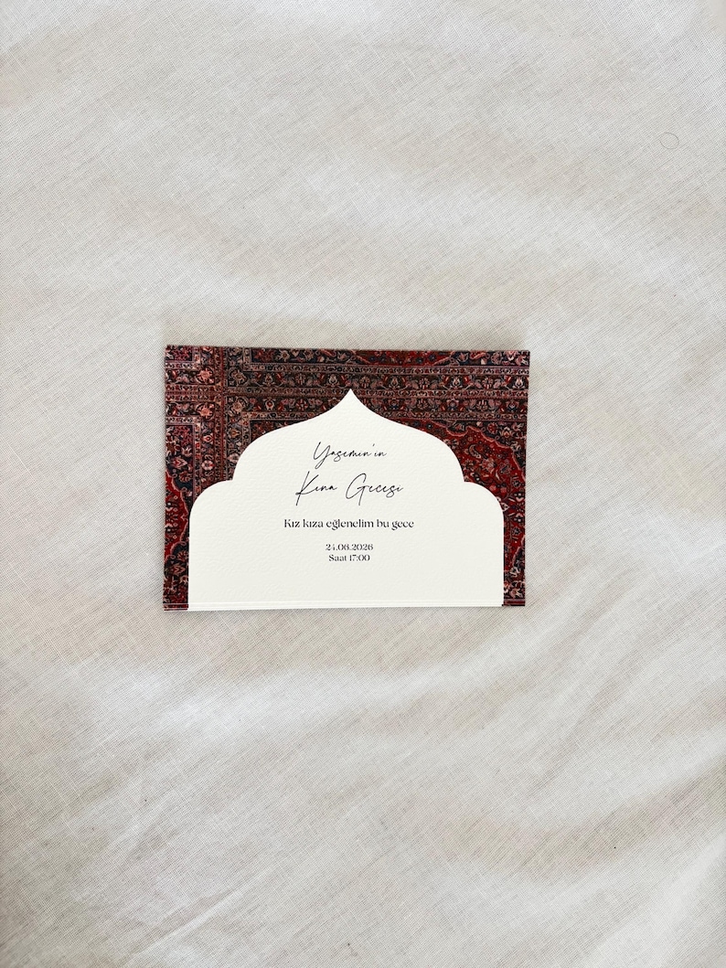 May include: A rectangular invitation card with a white cutout design and text. The card features a patterned red and brown background reminiscent of a rug. The text is in a script font.