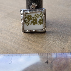 May include: Square pendant with a white background and green floral design. The pendant is framed in silver and hangs from a silver bail. The pendant is approximately 1 inch square.