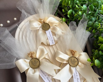 May include: Ivory-colored wedding favors with gold accents. Each favor features a cream-colored bow, a gold ring, and a small tag with text. The favors are arranged on a textured, off-white surface, with greenery in the background.