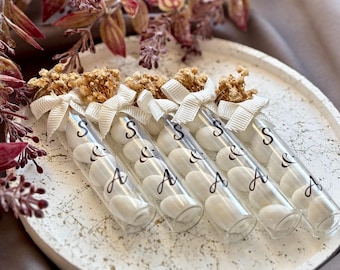 May include: A collection of clear glass tubes filled with white candies, arranged on a round, textured, white tray. Each tube is adorned with a white ribbon and dried flowers. The tubes are labeled with the letters "S & A".