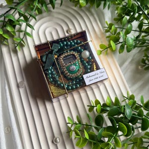 May include: A gift set featuring a green beaded prayer beads and a digital counter, presented in a clear box with a gold ribbon. The set includes a small card with the names "Muhammad & Nara" and the date "June 28th 2026". The set is on a white tray with green foliage.