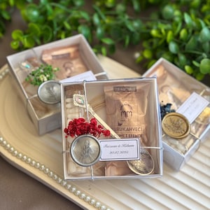 May include: Clear square boxes containing small gifts, tied with silver string. Each box includes a small bag of tea, a decorative spoon, and a wax seal. The boxes are arranged on a white tray with a pearl border. The text "Faizaan & Halum 20.07.2025" is visible.