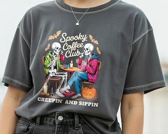 May include: Dark gray t-shirt featuring a Halloween-themed graphic. The design shows two skeletons at a table with coffee cups, the text "Spooky Coffee Club" and "CREEPIN' AND SIPPIN". A pumpkin and bats complete the spooky scene.