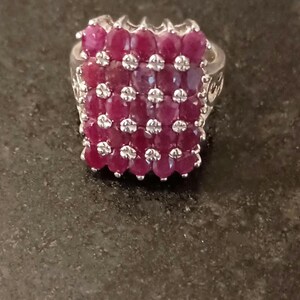 May include: A silver-toned ring featuring a rectangular cluster of multiple oval-cut deep red rubies arranged in a grid pattern, interspersed with small clear accent stones between the rubies, set on a polished metal band with decorative side details.