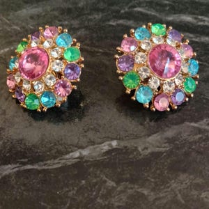 May include: A pair of round, gold-tone earrings with a cluster of colorful rhinestones. The earrings feature a large pink rhinestone in the center surrounded by smaller rhinestones in shades of green, blue, purple, and pink.