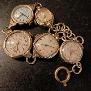 May include: A gold-tone charm bracelet featuring five vintage watch faces of various shapes and sizes. The watches include round and square cases with white dials, Arabic numerals, and metallic hands, all connected by a series of circular gold-tone links.
