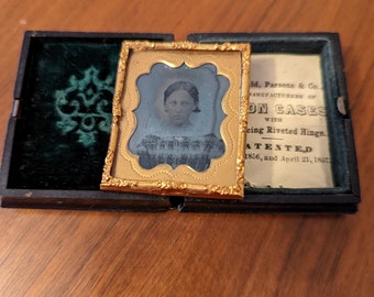 May include: A black and white photograph of a young woman in a gold-colored frame. The frame has a decorative border and is set against a green velvet background.