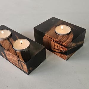 Könnte beinhalten: Zwei rechteckige Kerzenhalter aus dunklem Holz mit natürlicher Maserung. Jeder Halter hat eine Aussparung zur Aufnahme einer brennenden Teelichtkerze. Das Holz hat eine dunkle, fast schwarze Oberfläche.