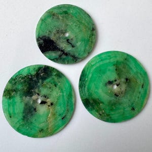 May include: Three circular, flat stone discs featuring a vibrant green color with natural black and white inclusions. Each disc has a small, centered circular hole, showcasing a polished surface with unique mineral patterns and variations.