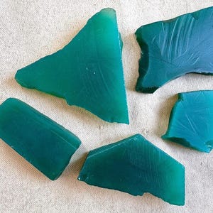 Könnte beinhalten: Fünf unregelmäßig geformte, durchscheinende, teal-farbene Glasstücke sind auf einer hellen, strukturierten Oberfläche angeordnet. Die Stücke variieren in Größe und Form, einige weisen subtile Oberflächenstrukturen und Unvollkommenheiten auf.