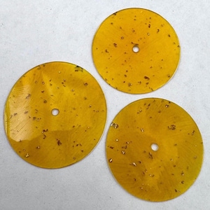 Pode incluir: Três discos circulares e planos de resina na cor âmbar, cada um com um orifício central e incrustado com pequenas partículas metálicas em tom dourado espalhadas pelo material translúcido, dispostos em formação triangular.