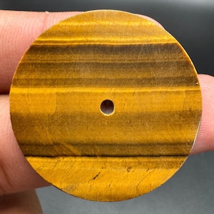 Pode incluir: Um cabochão circular de pedra olho-de-tigre com faixas horizontais de amarelo dourado e castanho escuro. A pedra tem uma superfície lisa e polida com um único pequeno orifício circular centrado perfurado no meio do disco.