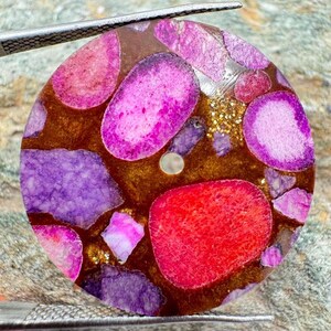 Op de afbeelding: Een ronde hanger met een mozaïek van levendige roze, paarse en rode steenfragmenten ingebed in een doorschijnende barnsteenkleurige harsbasis met gouden glitteraccenten. Het stuk heeft een klein centraal gat en wordt vastgehouden door een metalen pincet.