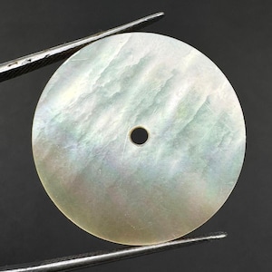 Könnte beinhalten: Eine kreisförmige Perlmutt-Scheibe mit einer irisierenden, perlmuttartigen Oberfläche in dezenten Weiß- und Hellgrüntönen. Die Scheibe hat ein kleines, mittiges Loch und wird von einer Metallpinzette gehalten, was ihre glatte Textur zeigt.