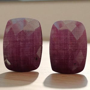 Könnte beinhalten: Zwei facettierte, oval geformte Edelsteine in einem tiefen Rot-Violett-Ton. Die Edelsteine haben eine polierte Oberfläche und werden auf einer hellen Holzoberfläche präsentiert. Der Schliff der Steine erzeugt mehrere reflektierende Facetten.