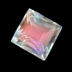 Könnte beinhalten: Ein quadratisch geschliffener Edelstein mit einem rosa Zentrum und schillernden Kanten. Der Edelstein ist facettiert und reflektiert das Licht, wobei eine Reihe von Farben angezeigt werden, darunter Rosa, Blau und Lila. Der Hintergrund ist tiefschwarz.