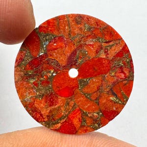 Pode incluir: Um pingente circular e plano com um mosaico vibrante de fragmentos de pedra vermelha e laranja, intercalados com veios de cor dourada metálica. A peça tem uma superfície lisa e polida com um pequeno orifício circular centralizado.