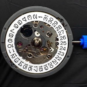 Op de afbeelding: Een rond mechanisch horloge-uurwerk met een witte datumschijf met zwarte cijfers van 1 tot 31. Het metalen interne mechanisme toont tandwielen, schroeven en kleine rode robijnen. Een blauwe plastic stift steekt uit de zijkant van het geheel.