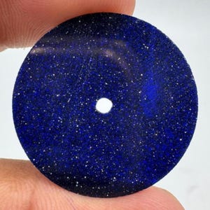 Pode incluir: Um pingente circular e plano de pedra do sol azul, apresentando uma cor de base azul cobalto profunda com inúmeras partículas metálicas incrustadas que criam um efeito cintilante semelhante a estrelas. Possui um pequeno orifício central.