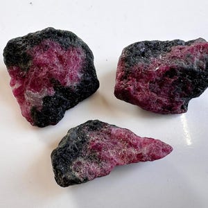Könnte beinhalten: Drei rohe Rubinsteine mit einer dunklen, fast schwarzen Außenschicht und leuchtend rosa-roten Innenräumen. Die rohen Edelsteine werden vor einem weißen Hintergrund präsentiert und zeigen ihre natürlichen Texturen und Farben.