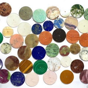 Può includere: Una collezione di cabochon rotondi in pietre preziose di vari colori e motivi. Le pietre preziose includono malachite, occhio di tigre, lapislazzuli e altre. Ogni cabochon ha un piccolo foro al centro.