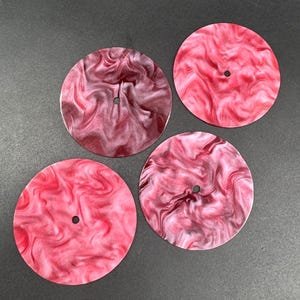May include: Four round coasters with a swirled, marbled design in shades of red and burgundy. Each coaster has a small hole in the center. The coasters are arranged on a dark gray surface.