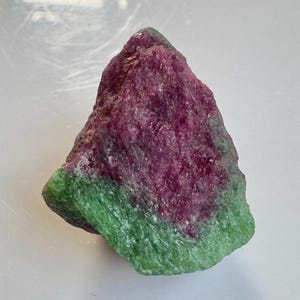 Könnte beinhalten: Ein rohes Rubin- und Zoisit-Exemplar. Der Stein weist eine lebendige Farbkombination auf, wobei ein tiefes Magenta den oberen Teil dominiert und ein auffälliges Grün die Basis bildet. Die natürliche Mineralformation hat eine strukturierte Oberfläche.