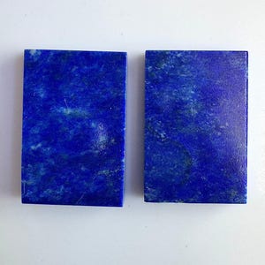 Puede incluir: Dos piedras rectangulares de lapislázuli. Las piedras son de color azul profundo con motas doradas y blancas. Las piedras son lisas y pulidas, y parecen listas para su uso en joyería u otras manualidades.
