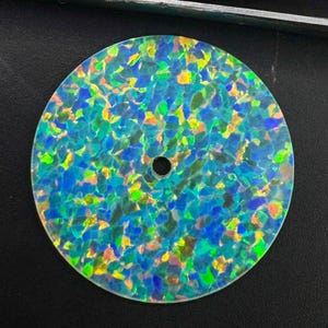Könnte beinhalten: Eine kreisförmige, flache Scheibe mit einem lebendigen Mosaikmuster aus irisierenden blauen, grünen und gelben Flecken. Die Oberfläche weist ein Hochglanz-Finish mit einem kleinen, mittig platzierten kreisförmigen Loch auf, das ein reflektierendes Erscheinungsbild erzeugt.