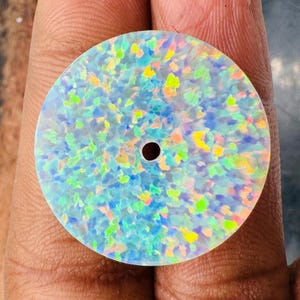 May include: A round, iridescent opal stone with a central hole. The opal displays a mosaic of colors, including blue, green, yellow, and orange, set against a white background. The stone is held in a hand, showcasing its polished surface.