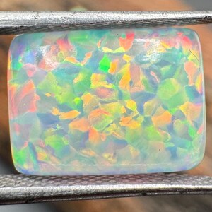 Könnte beinhalten: Ein rechteckiger Opal-Edelstein, gehalten von einer Metallpinzette, zeigt ein lebendiges Farbspiel, darunter Grün, Orange und Gelb. Die polierte Oberfläche des Edelsteins wirkt durchscheinend und zeigt sein inneres Farbspiel.