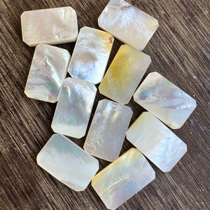 Könnte beinhalten: Eine Sammlung rechteckiger Perlmuttperlen mit schillernden Oberflächen. Die Perlen haben abgerundete Ecken und variieren in der Farbe von Weiß bis Hellgelb. Sie sind auf einer dunkelbraunen Holzoberfläche angeordnet.