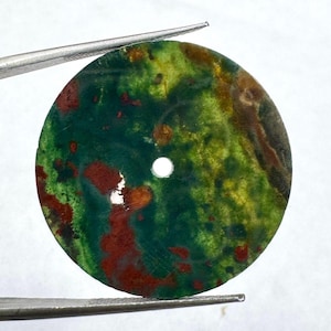 May include: A circular, flat stone disc featuring a mottled pattern of deep forest green, vibrant lime green, and earthy reddish-brown patches. The disc has a small, centred circular hole and is held by metal tweezers against a white background.