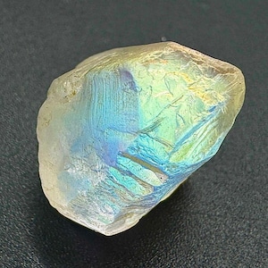 Könnte beinhalten: Ein grob geschliffener, schillernder Edelstein mit hellgelber Basis und Blitzen von Blau, Grün und Lila. Die Oberfläche hat ein strukturiertes Aussehen und reflektiert das Licht in einer schimmernden Darstellung. Der Edelstein ist wahrscheinlich ein Mineralexemplar.