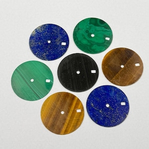 Könnte beinhalten: Eine Sammlung von sieben kreisförmigen Zifferblättern. Die Zifferblätter bestehen aus verschiedenen Materialien, darunter blauer Lapislazuli, grüner Malachit, schwarzes Holz und Tigerauge. Jedes Zifferblatt hat ein kleines Loch und einen rechteckigen Ausschnitt.