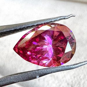 Könnte beinhalten: Ein tropfenförmiger, facettierter Edelstein mit leuchtend rosa Farbton, gehalten von einer silbernen Pinzette. Die innere Struktur des Edelsteins reflektiert das Licht und erzeugt ein schillerndes Farb- und Brillanzspiel. Der Edelstein ist vor weißem Hintergrund.