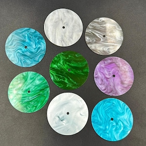 May include: Nine round watch faces in various colors, including teal, green, purple, and silver. Each face has a swirling, marbled pattern and a small hole in the center. The faces are arranged on a dark surface.