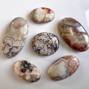 Puede incluir: Colección de seis piedras de ágata pulidas de diversas formas y tamaños. Las piedras exhiben una gama de colores, incluyendo beige, marrón, rojo y gris, con intrincados patrones y bandas. Las piedras están dispuestas sobre una superficie blanca.