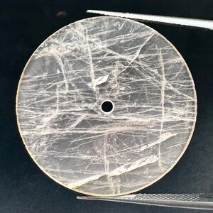 May include: A translucent, round gemstone with a central hole and visible internal fractures. The stone is held by tweezers, showcasing its delicate, light color and intricate patterns. The gemstone is likely for jewelry making.
