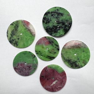 Op de afbeelding: Zes ronde, gepolijste edelsteen wijzerplaten. De wijzerplaten tonen een mix van groene, zwarte en rode kleuren. Elke wijzerplaat heeft een klein gaatje in het midden. De edelstenen hebben een natuurlijk, gemarmerd uiterlijk.