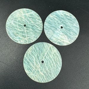 May include: Three circular, pale blue-green stone discs, each with a central hole. The stone features a natural, textured pattern with white and darker green streaks. The discs are displayed on a dark surface.