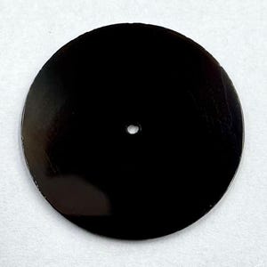 Pode incluir: Um objeto circular preto brilhante com um pequeno orifício no centro. A superfície reflete a luz, sugerindo um material sólido. É provavelmente um componente ou parte de um produto maior.