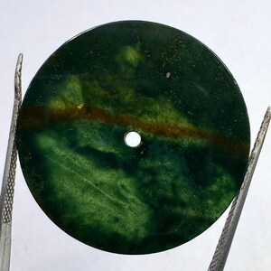 Puede incluir: Una piedra preciosa redonda y pulida con un agujero, sujeta con pinzas. La piedra presenta una mezcla de tonos verde oscuro y marrón, con un efecto veteado verde más claro. La piedra tiene aproximadamente 3,8 cm de diámetro.