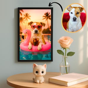 May include: A framed print depicts a dog wearing heart-shaped sunglasses and a pink flamingo float, enjoying a drink in a pool. The background shows palm trees and a sunset. A small cat figurine, a rose in a vase, and a book are on a table.